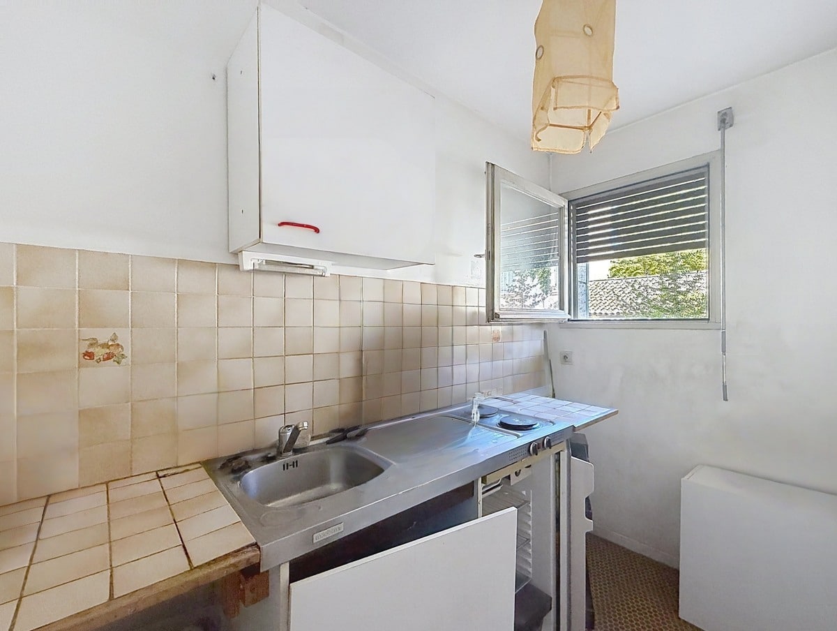 Cuisine étroite avec carrelage mural beige, évier en inox double bac et plaque de cuisson intégrée sur plan de travail carrelé, associés à un réfrigérateur blanc visible sur le côté. Une fenêtre à jalousies métalliques laisse entrer la lumière naturelle sur l'espace cuisine, au-dessus duquel est fixé un meuble haut blanc.