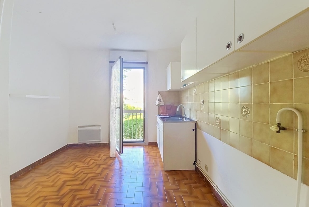 Cuisine indépendante avec un sol en tomettes rouges en chevrons, des meubles hauts blancs et un carrelage mural beige décoré, dotée d'un évier encastré sous fenêtre et d'un convecteur électrique mural. Une porte-fenêtre ouverte donne accès à un balcon avec vue sur des arbres verts.
