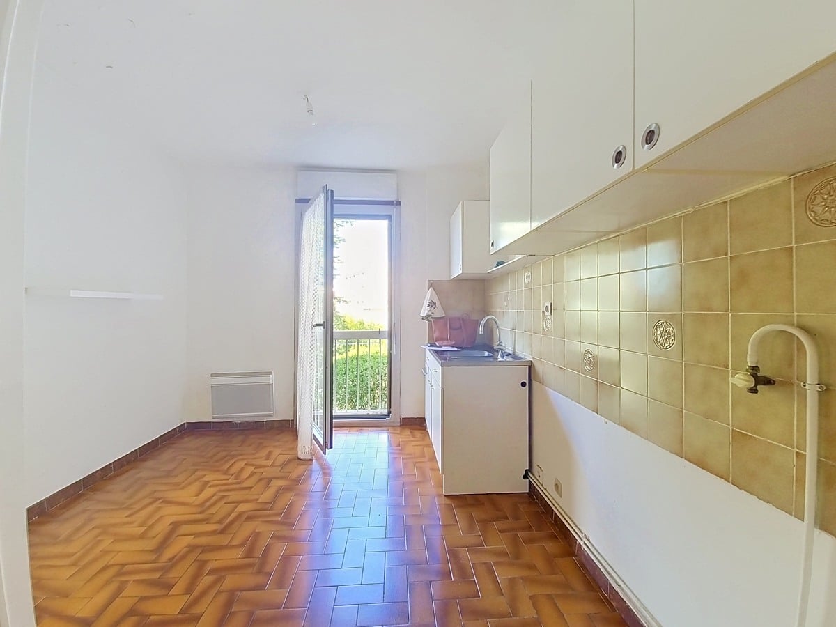 Cuisine indépendante avec un sol en tomettes rouges en chevrons, des meubles hauts blancs et un carrelage mural beige décoré, dotée d'un évier encastré sous fenêtre et d'un convecteur électrique mural. Une porte-fenêtre ouverte donne accès à un balcon avec vue sur des arbres verts.