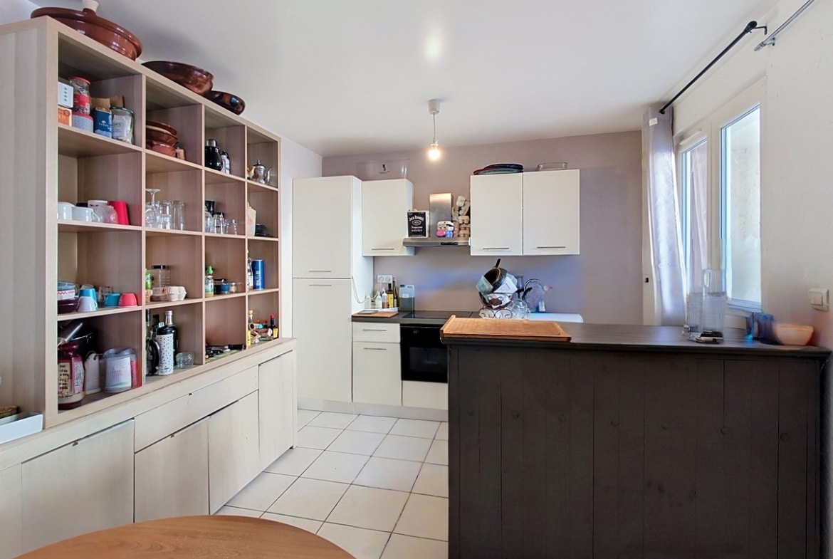 Cuisine indépendante avec bibliothèque ouverte en bois clair intégrée sur toute la hauteur, chargée de vaisselle, bocaux et ustensiles variés. Îlot central en bois foncé avec plan de travail sombre, armoires hautes blanches et fenêtre avec rideau blanc côté droit.