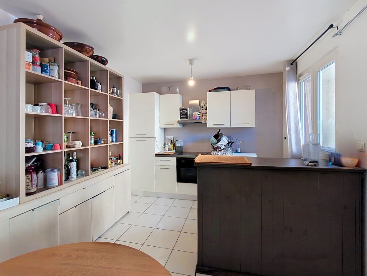 Cuisine indépendante avec bibliothèque ouverte en bois clair intégrée sur toute la hauteur, chargée de vaisselle, bocaux et ustensiles variés. Îlot central en bois foncé avec plan de travail sombre, armoires hautes blanches et fenêtre avec rideau blanc côté droit.