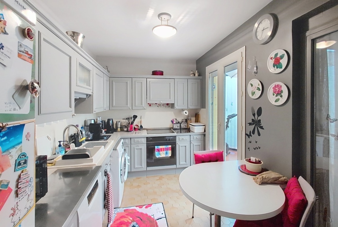 Cuisine en longueur aux meubles gris clair de style classique avec plan de travail blanc, évier inox et électroménager encastré dont un four noir et un lave-linge. Un coin repas avec table ronde blanche et banquette rouge est installé côté droit, près d'une porte-fenêtre donnant sur l'extérieur.