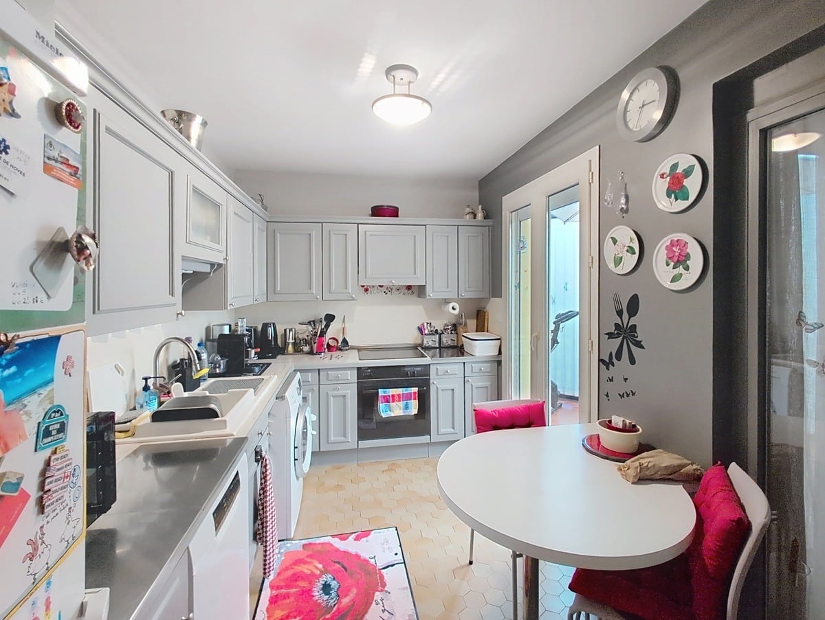 Cuisine en longueur aux meubles gris clair de style classique avec plan de travail blanc, évier inox et électroménager encastré dont un four noir et un lave-linge. Un coin repas avec table ronde blanche et banquette rouge est installé côté droit, près d'une porte-fenêtre donnant sur l'extérieur.