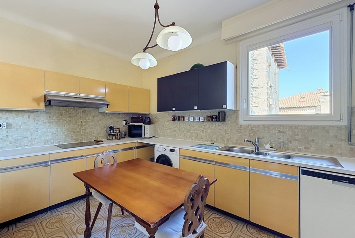 Cuisine en L avec façades de placards jaune moutarde et plans de travail en inox, équipée d'une table de cuisson, d'un lave-linge encastré, d'un lave-vaisselle Siemens et d'une hotte aspirante, avec un carrelage de sol à motifs floraux en ciment. Une fenêtre à double vitrage offre une vue sur les toits en tuiles d'un bâtiment en pierre voisin.