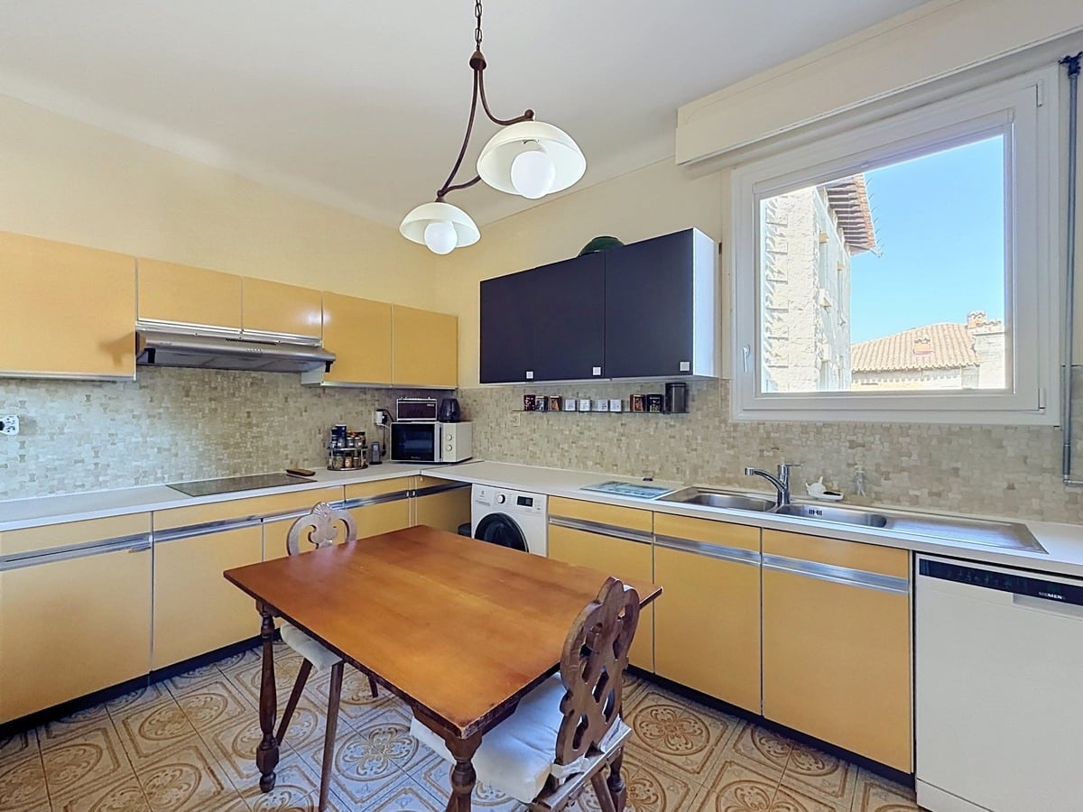 Cuisine en L avec façades de placards jaune moutarde et plans de travail en inox, équipée d'une table de cuisson, d'un lave-linge encastré, d'un lave-vaisselle Siemens et d'une hotte aspirante, avec un carrelage de sol à motifs floraux en ciment. Une fenêtre à double vitrage offre une vue sur les toits en tuiles d'un bâtiment en pierre voisin.