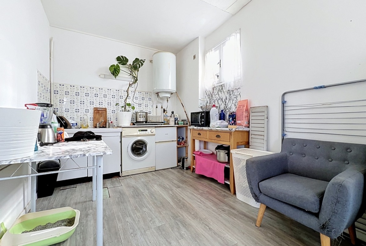 Cuisine en L avec plan de travail bas, équipée d'un lave-linge encastré, d'une plaque de cuisson électrique, d'un four micro-ondes et d'un chauffe-eau ballon mural blanc cylindrique en hauteur. Crédence en faïence à motifs bleus, sol en revêtement imitation parquet clair, fauteuil gris capitonné à pieds bois et bac à litière vert au premier plan.