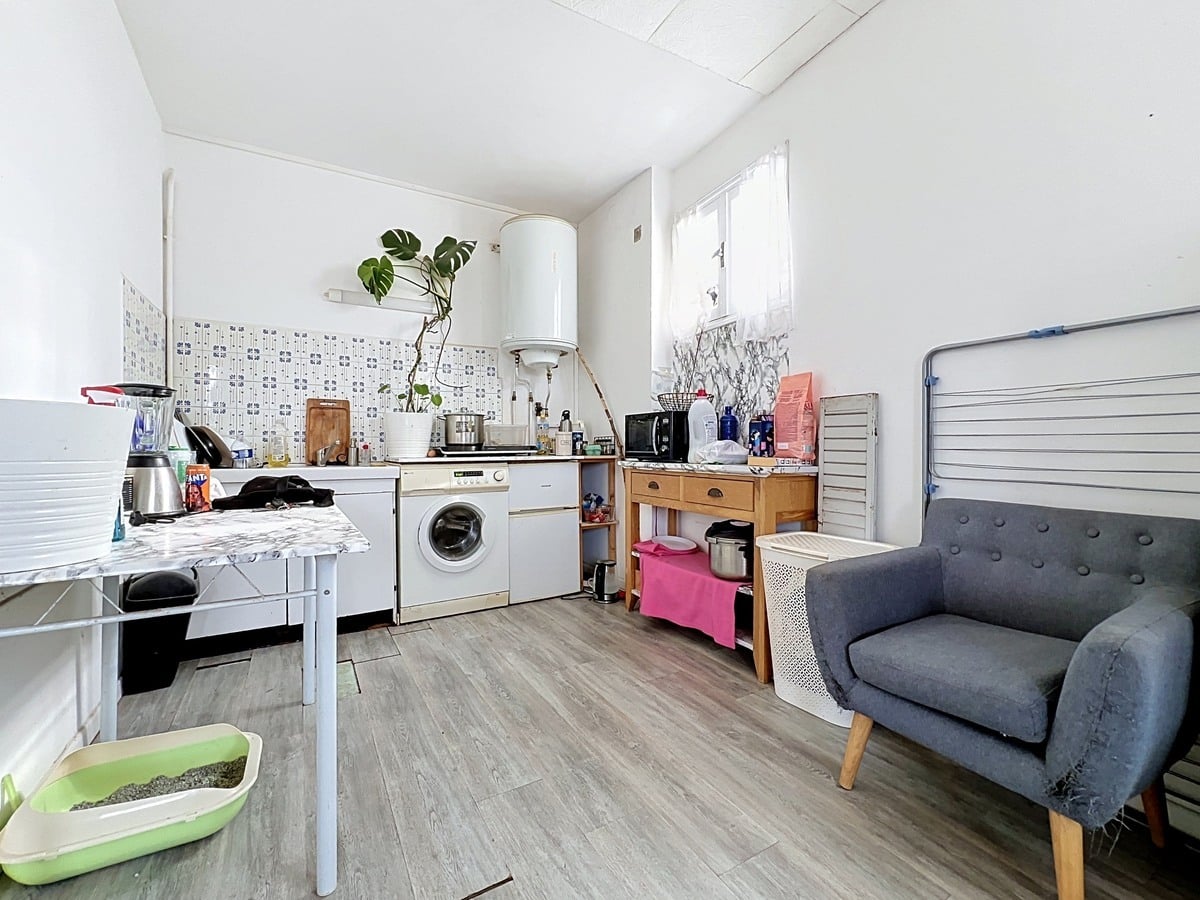Cuisine en L avec plan de travail bas, équipée d'un lave-linge encastré, d'une plaque de cuisson électrique, d'un four micro-ondes et d'un chauffe-eau ballon mural blanc cylindrique en hauteur. Crédence en faïence à motifs bleus, sol en revêtement imitation parquet clair, fauteuil gris capitonné à pieds bois et bac à litière vert au premier plan.