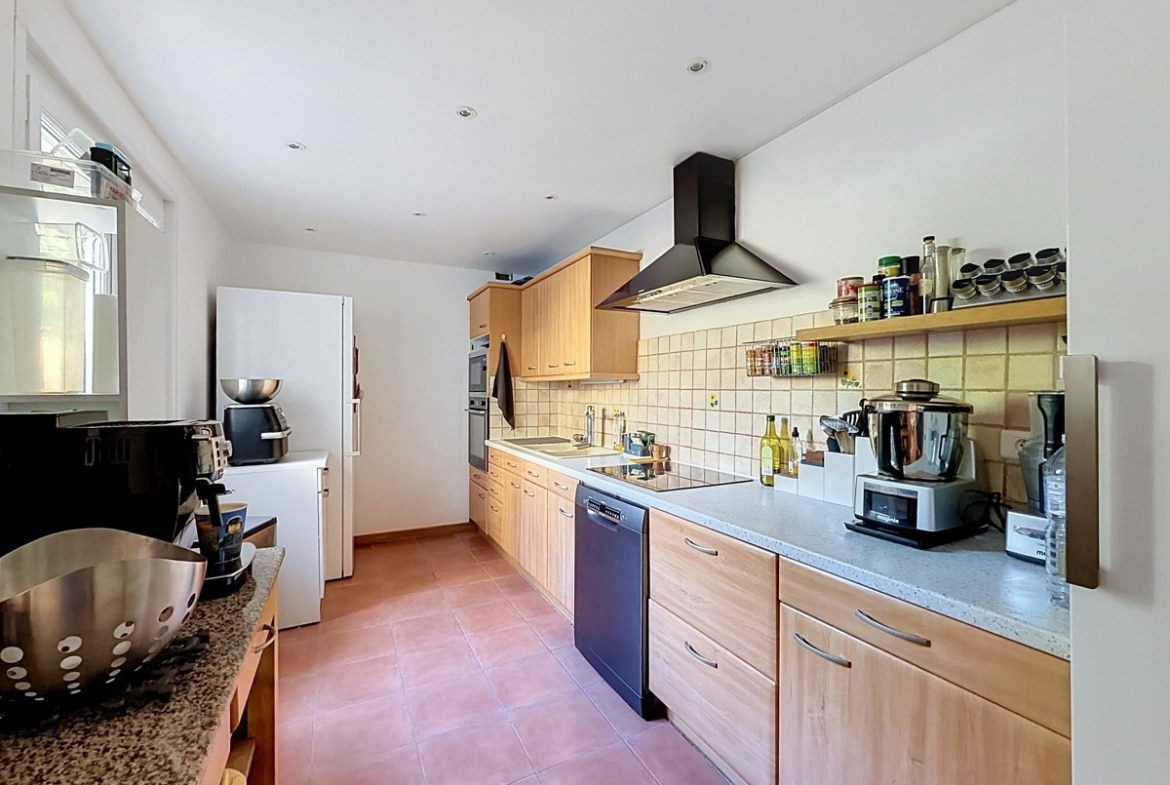 Cuisine en L avec façades en bois clair, plan de travail gris et carrelage mural beige clair. Lave-vaisselle encastré, hotte aspirante noire, réfrigérateur américain blanc et nombreux appareils électroménagers posés sur le plan de travail.