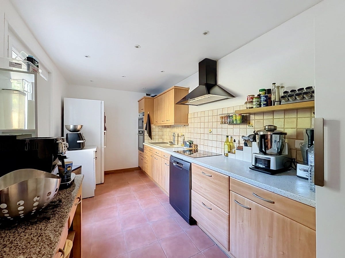 Cuisine en L avec façades en bois clair, plan de travail gris et carrelage mural beige clair. Lave-vaisselle encastré, hotte aspirante noire, réfrigérateur américain blanc et nombreux appareils électroménagers posés sur le plan de travail.