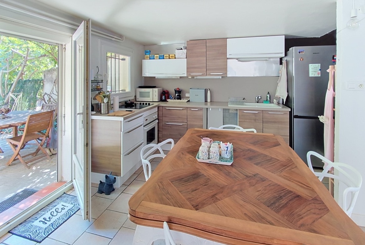 Cuisine en L avec façades en bois clair et blanc mat, équipée d'un réfrigérateur inox, d'un four encastré et d'une plaque de cuisson, avec un îlot central en bois massif à motif chevron entouré de chaises blanches. Une porte-fenêtre à double vantail ouvre directement sur la terrasse extérieure avec salon de jardin en teck.