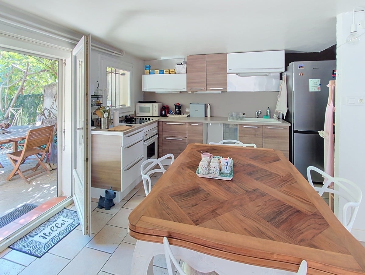 Cuisine en L avec façades en bois clair et blanc mat, équipée d'un réfrigérateur inox, d'un four encastré et d'une plaque de cuisson, avec un îlot central en bois massif à motif chevron entouré de chaises blanches. Une porte-fenêtre à double vantail ouvre directement sur la terrasse extérieure avec salon de jardin en teck.