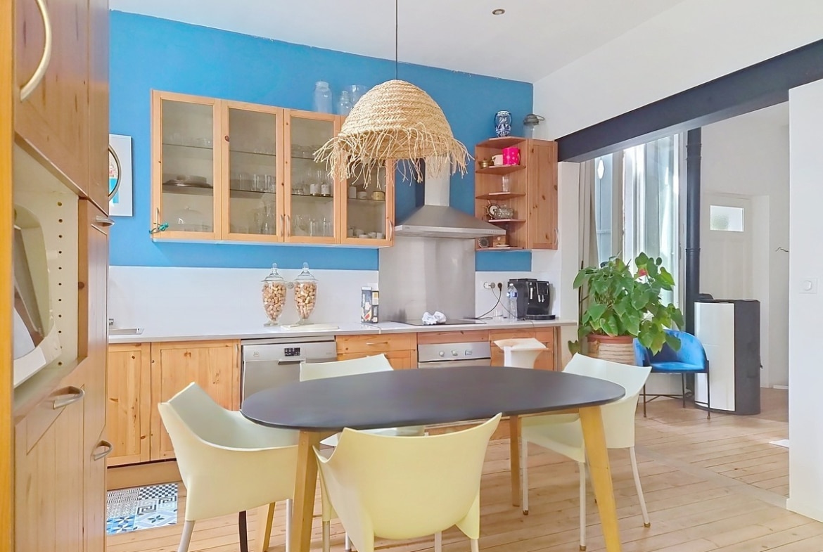 Cuisine équipée avec façade de meubles en pin, hotte aspirante murale et crédence blanche sur fond de mur peint en bleu vif. Table ovale noire entourée de chaises blanches et jaunes sous un luminaire en fibre naturelle tressée, parquet bois clair au sol.