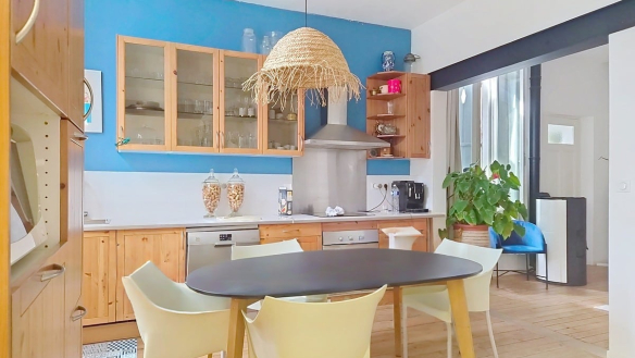 Cuisine équipée avec façade de meubles en pin, hotte aspirante murale et crédence blanche sur fond de mur peint en bleu vif. Table ovale noire entourée de chaises blanches et jaunes sous un luminaire en fibre naturelle tressée, parquet bois clair au sol.