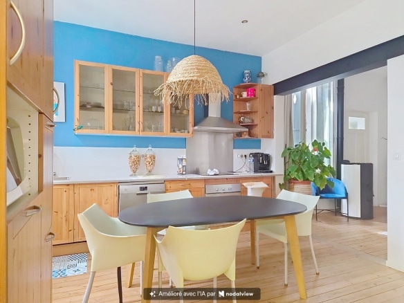 Cuisine équipée avec façade de meubles en pin, hotte aspirante murale et crédence blanche sur fond de mur peint en bleu vif. Table ovale noire entourée de chaises blanches et jaunes sous un luminaire en fibre naturelle tressée, parquet bois clair au sol.