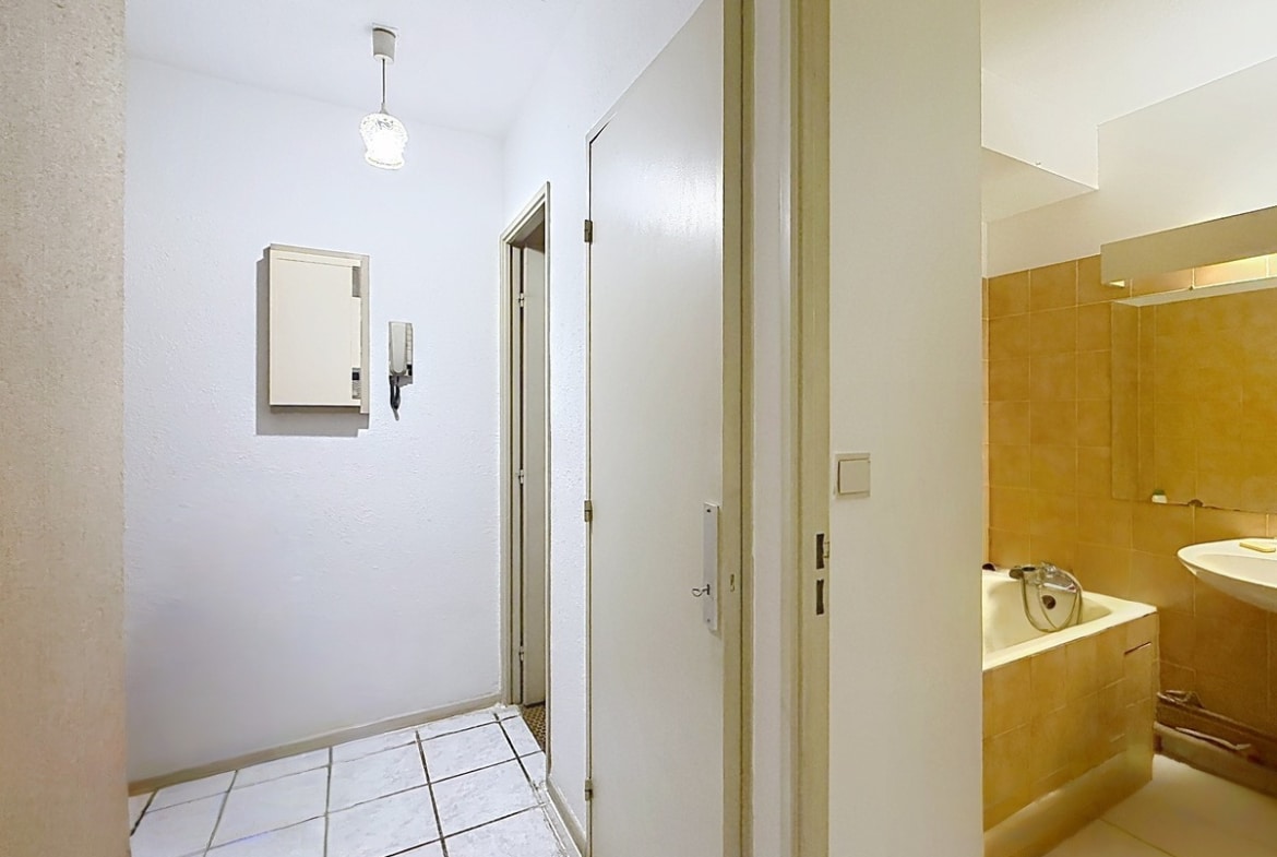 Couloir d'entrée carrelé en blanc avec interphone mural et boîtier électrique encastré, donnant accès à plusieurs portes blanches desservant les pièces de l'appartement. Sur la droite, la salle de bain est partiellement visible avec son carrelage mural jaune ocre, sa baignoire encastrée et son lavabo blanc.