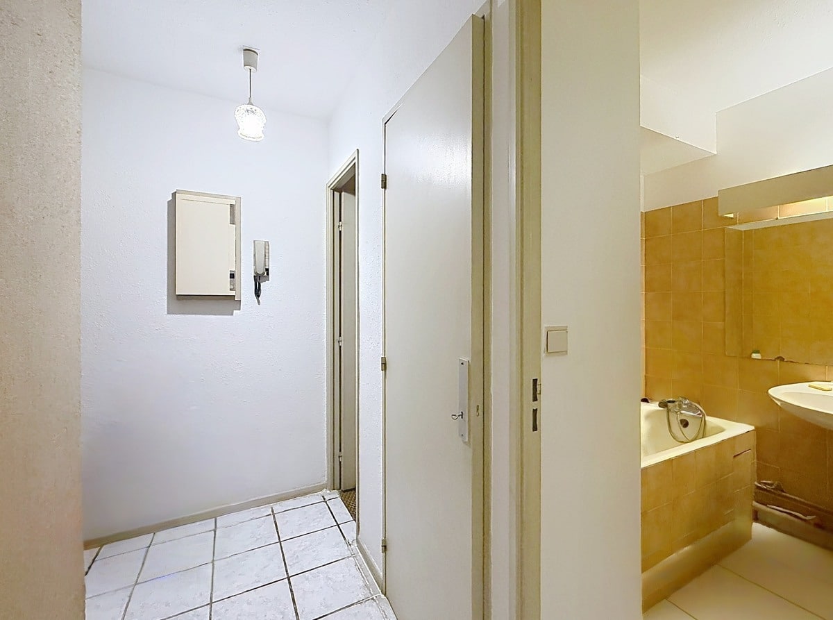 Couloir d'entrée carrelé en blanc avec interphone mural et boîtier électrique encastré, donnant accès à plusieurs portes blanches desservant les pièces de l'appartement. Sur la droite, la salle de bain est partiellement visible avec son carrelage mural jaune ocre, sa baignoire encastrée et son lavabo blanc.