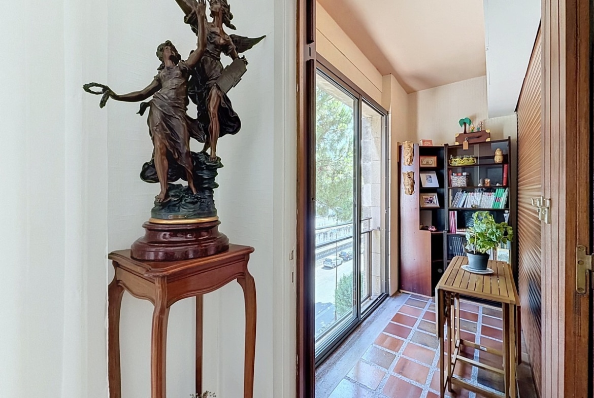 Couloir d'entrée avec une statue en bronze représentant deux figures féminines ailées posée sur un piédestal en bois sculpté, donnant accès à une loggia fermée par une baie vitrée coulissante avec vue sur la rue. La loggia est aménagée avec une étagère en bois sombre, un petit bureau pliant et un carrelage en terre cuite.
