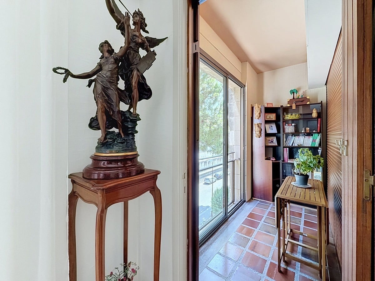 Couloir d'entrée avec une statue en bronze représentant deux figures féminines ailées posée sur un piédestal en bois sculpté, donnant accès à une loggia fermée par une baie vitrée coulissante avec vue sur la rue. La loggia est aménagée avec une étagère en bois sombre, un petit bureau pliant et un carrelage en terre cuite.