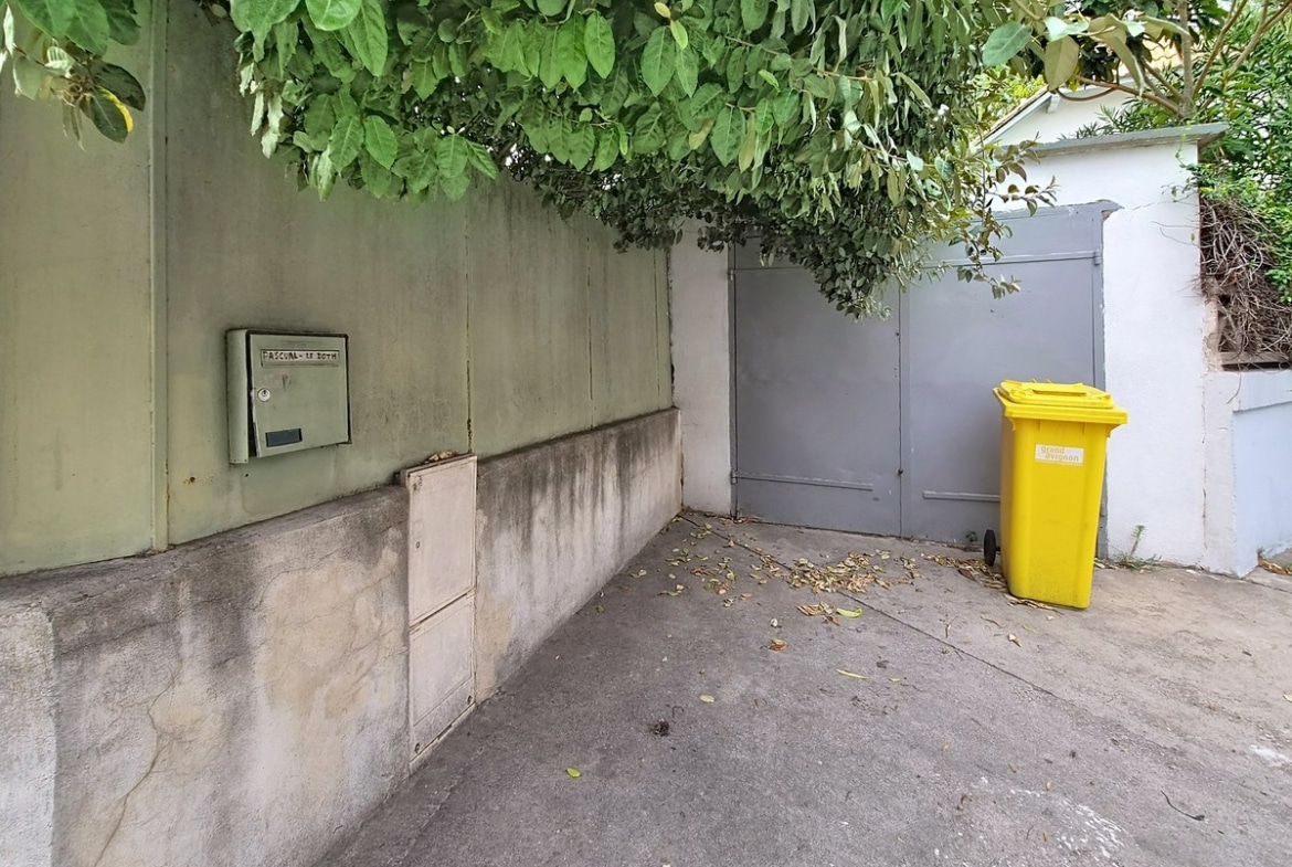 Entrée de propriété avec mur d'enceinte en béton enduit vert pâle, portail double battant en métal gris et boîte aux lettres encastrée métallique. Sol en béton gris avec feuilles mortes éparpillées, végétation dense en surplomb et bac de recyclage jaune positionné devant le portail.