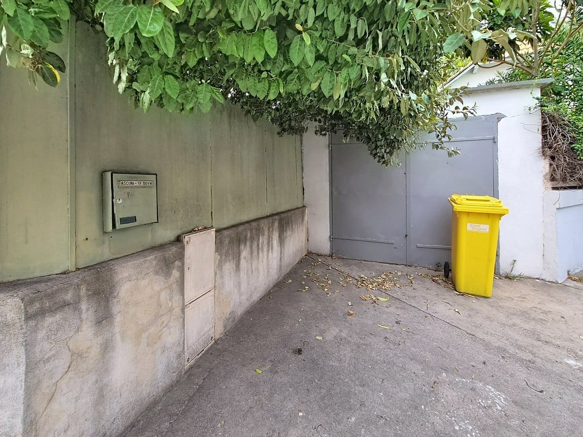 Entrée de propriété avec mur d'enceinte en béton enduit vert pâle, portail double battant en métal gris et boîte aux lettres encastrée métallique. Sol en béton gris avec feuilles mortes éparpillées, végétation dense en surplomb et bac de recyclage jaune positionné devant le portail.