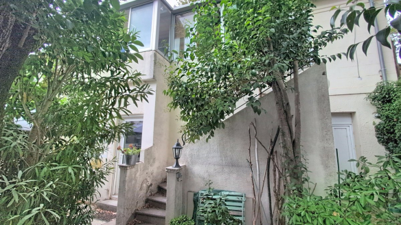 Façade en enduit clair d'une maison de deux niveaux, avec un escalier extérieur en béton menant à une entrée du rez-de-chaussée, bordé d'une rampe métallique et d'un lampadaire de jardin noir. Le premier étage présente une grande baie vitrée avec menuiserie aluminium ; la végétation abondante, dont bambous et arbustes, entoure la façade côté jardin.