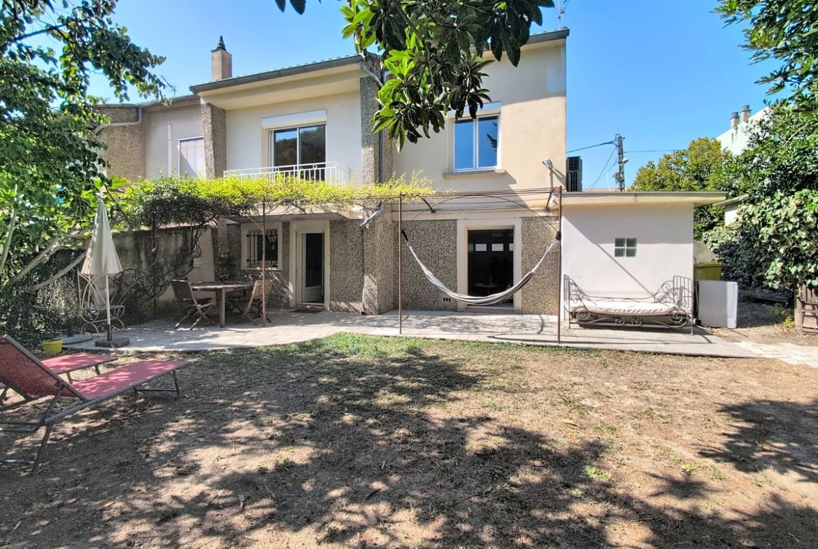 Maison de deux étages à enduit granité gris, dotée d'un balcon au premier étage avec garde-corps en fer forgé et d'une pergola métallique couverte de plantes grimpantes au rez-de-chaussée. Le jardin arboré comprend une terrasse dallée, un hamac, un bain de soleil rouge et un cabanon blanc sur la droite.