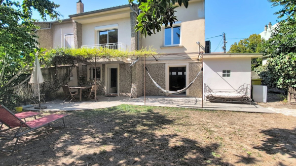 Maison de deux étages à enduit granité gris, dotée d'un balcon au premier étage avec garde-corps en fer forgé et d'une pergola métallique couverte de plantes grimpantes au rez-de-chaussée. Le jardin arboré comprend une terrasse dallée, un hamac, un bain de soleil rouge et un cabanon blanc sur la droite.