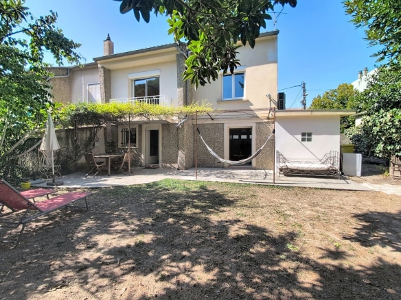 Maison de deux étages à enduit granité gris, dotée d'un balcon au premier étage avec garde-corps en fer forgé et d'une pergola métallique couverte de plantes grimpantes au rez-de-chaussée. Le jardin arboré comprend une terrasse dallée, un hamac, un bain de soleil rouge et un cabanon blanc sur la droite.