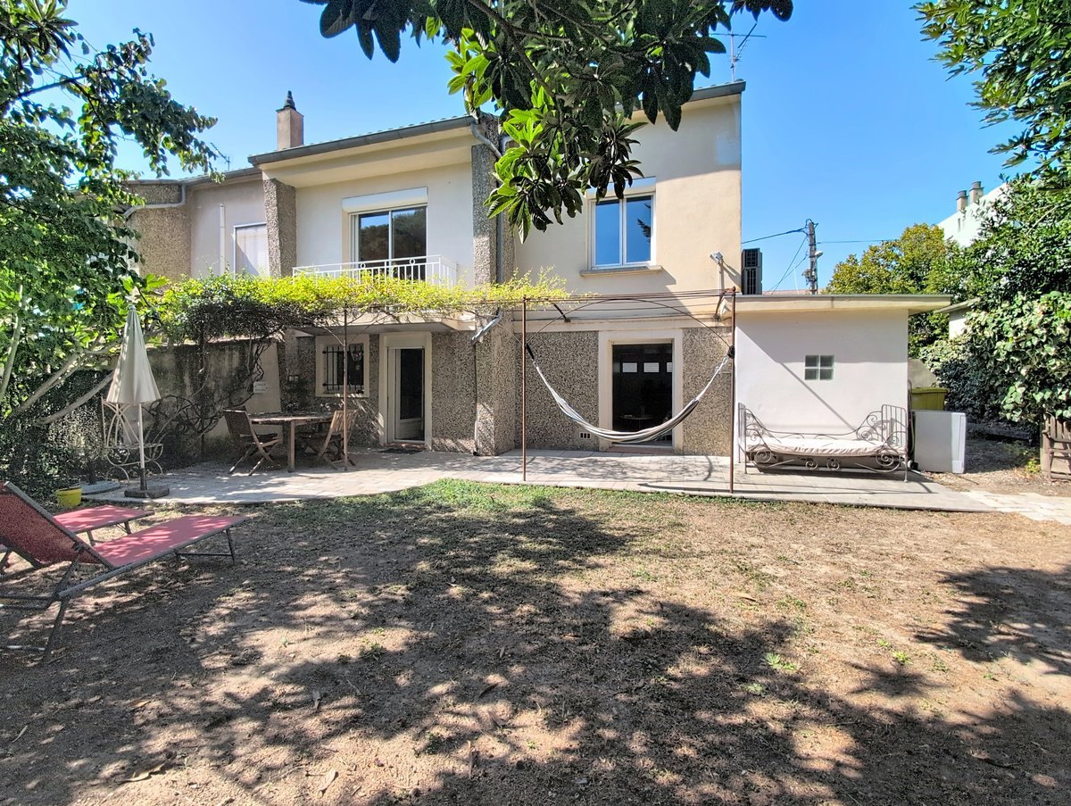 Maison de deux étages à enduit granité gris, dotée d'un balcon au premier étage avec garde-corps en fer forgé et d'une pergola métallique couverte de plantes grimpantes au rez-de-chaussée. Le jardin arboré comprend une terrasse dallée, un hamac, un bain de soleil rouge et un cabanon blanc sur la droite.