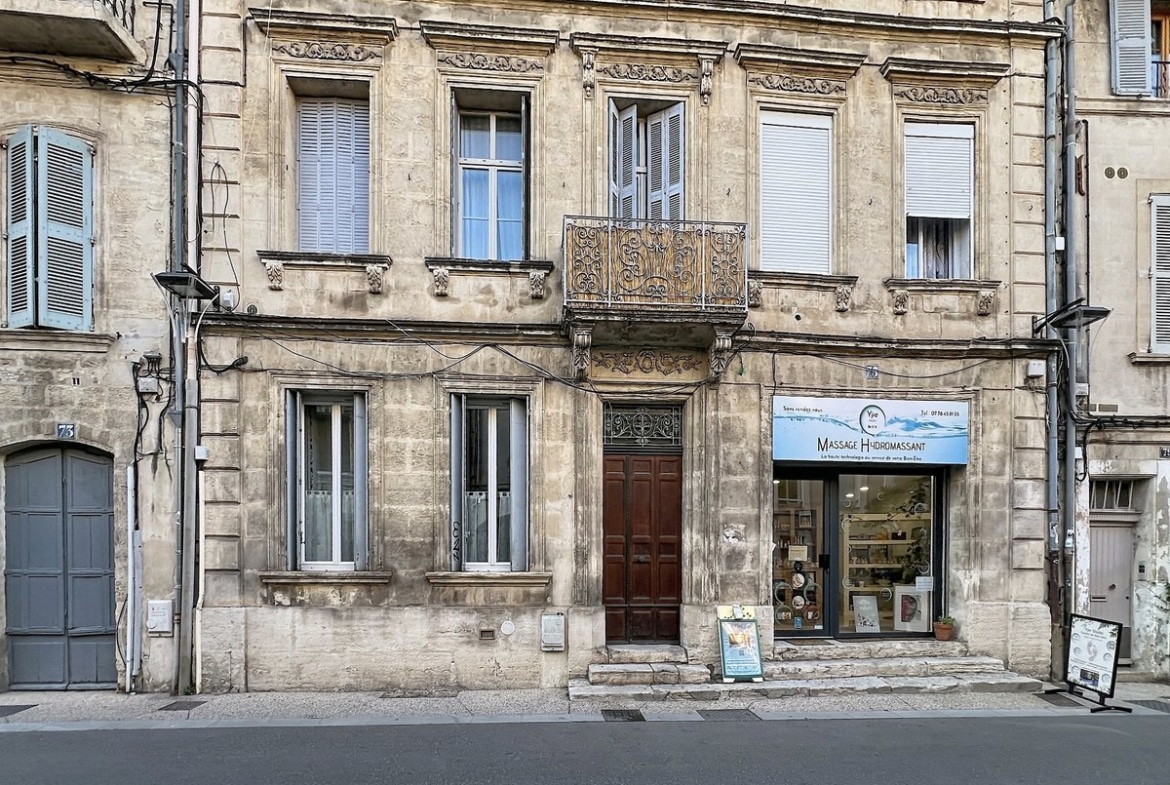 Façade haussmannienne en pierre de taille calcaire avec moulures sculptées, balcon en fer forgé ouvragé au premier étage et porte cochère en bois sombre à double vantail. Au rez-de-chaussée côté droit, devanture commerciale d'un salon de massage hydromassant avec vitrine et présentoirs visibles, numéro de rue 75 visible sur le pilastre gauche.