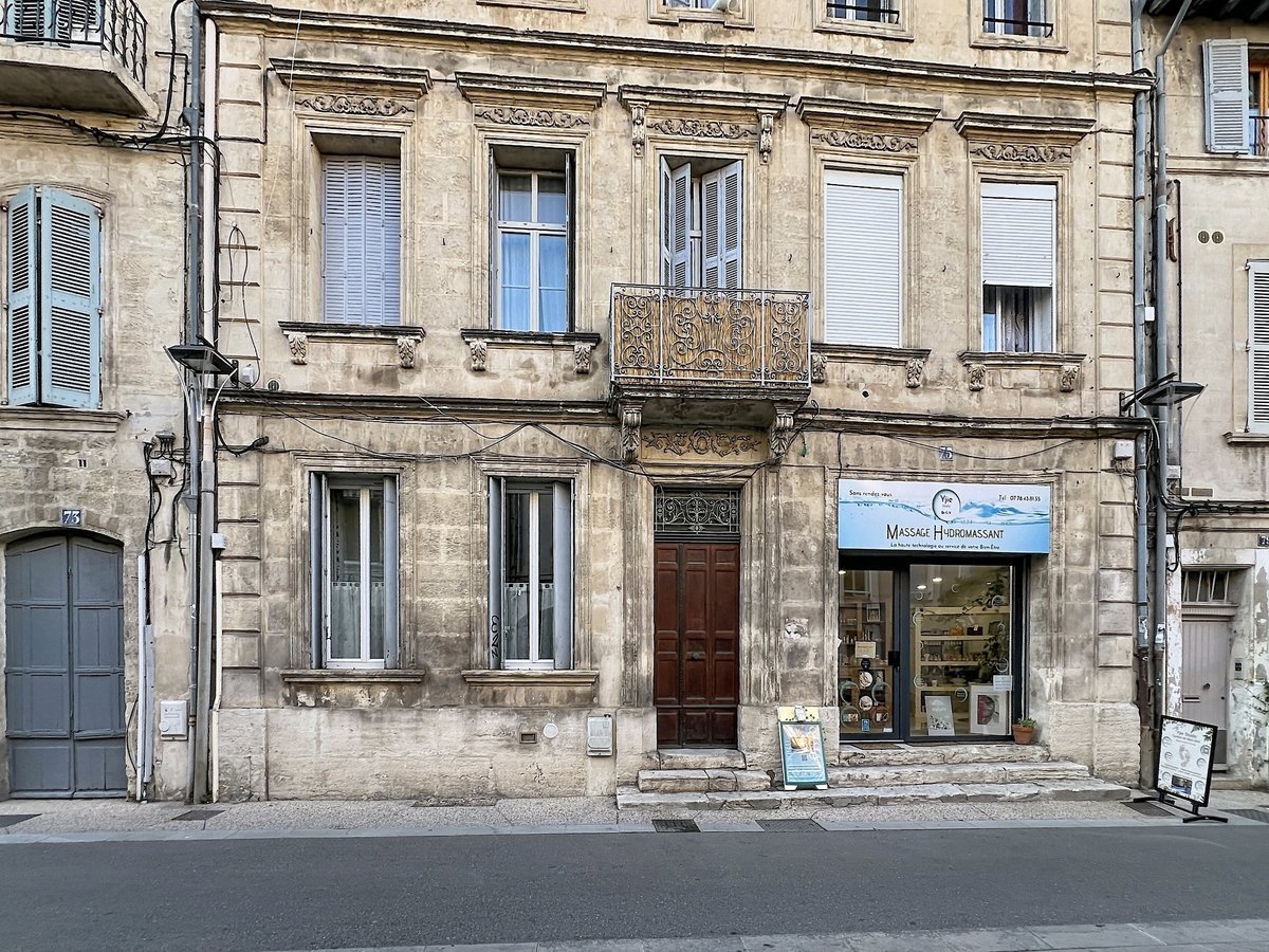 Façade haussmannienne en pierre de taille calcaire avec moulures sculptées, balcon en fer forgé ouvragé au premier étage et porte cochère en bois sombre à double vantail. Au rez-de-chaussée côté droit, devanture commerciale d'un salon de massage hydromassant avec vitrine et présentoirs visibles, numéro de rue 75 visible sur le pilastre gauche.