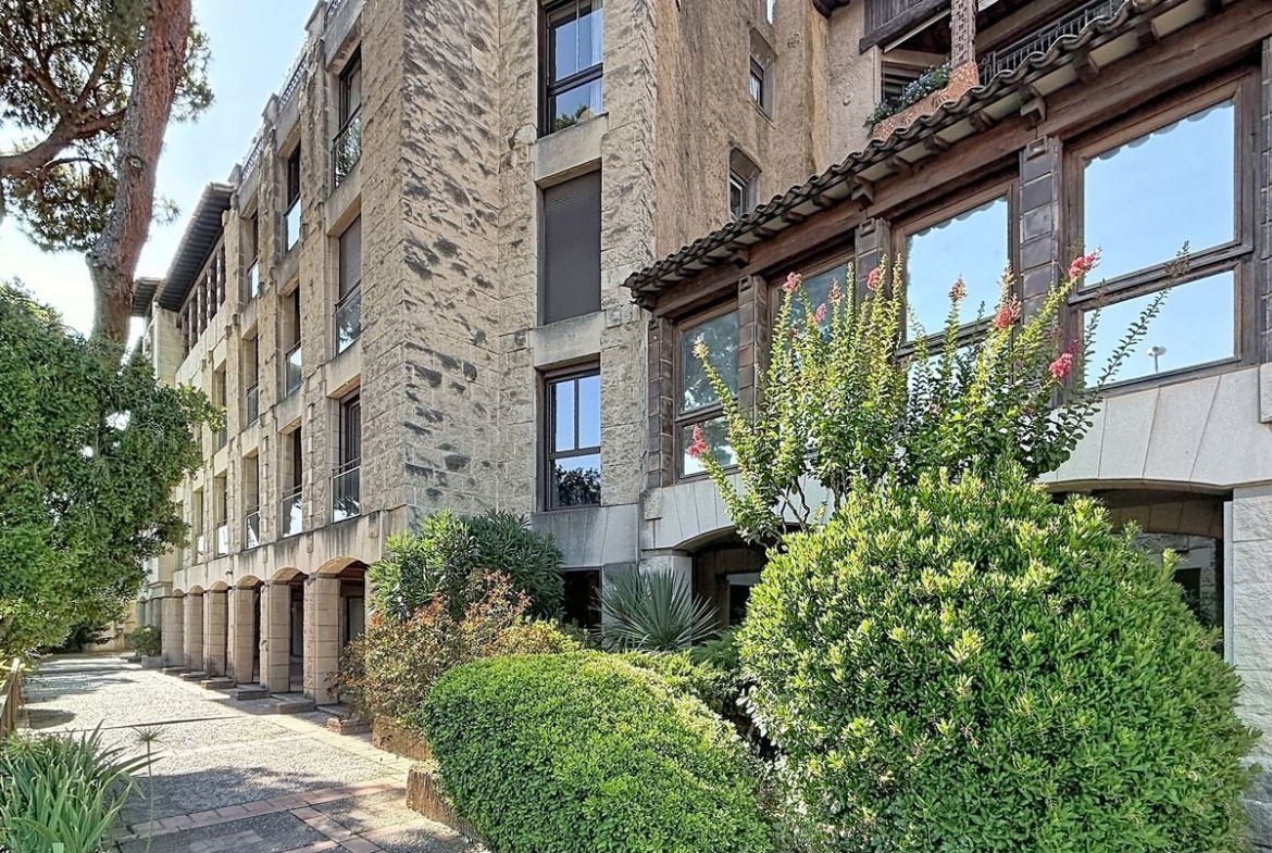 Façade d'un immeuble de quatre étages en pierre de taille beige avec des détails architecturaux en bois sombre, des arches au rez-de-chaussée et une toiture avec tuiles romaines aux accents méditerranéens, longée par une allée piétonne bordée d'arbustes taillés et de plantes fleuries roses. Le style architectural évoque une inspiration néo-régionaliste avec des bow-windows à ossature bois.