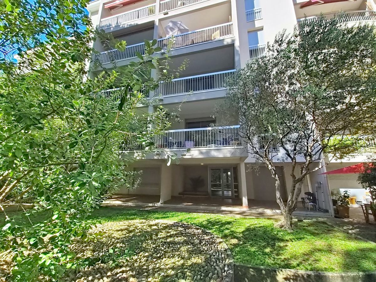 Immeuble résidentiel de cinq étages avec des balcons filants à garde-corps métalliques gris et un rez-de-chaussée en retrait formant un porche vitré, entouré d'un jardin planté d'oliviers, de buissons et d'une pelouse. La façade est enduite de blanc avec des tons beige, et de la végétation méditerranéenne occupe le premier plan.