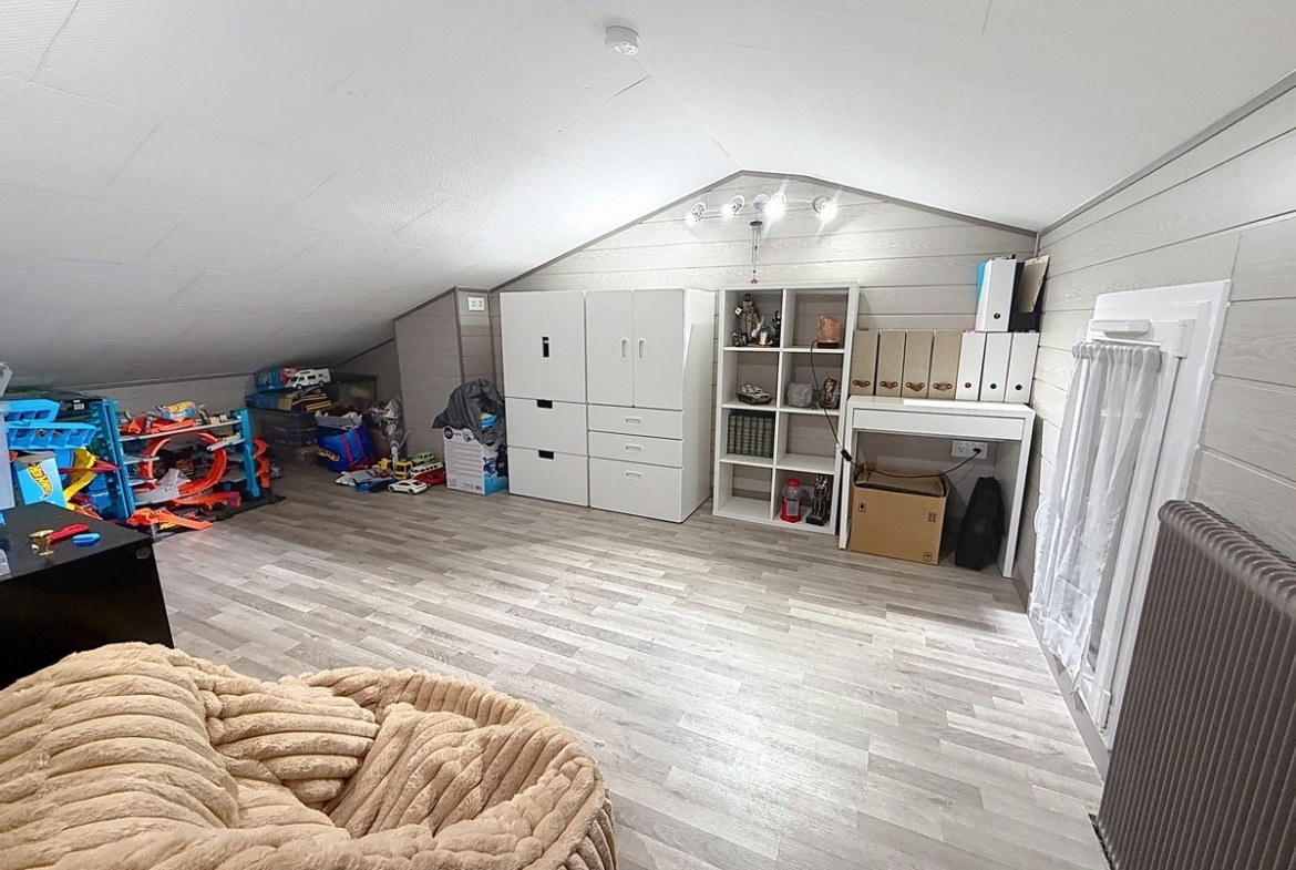Pièce sous combles aménagée avec sol en parquet stratifié gris clair et plafond incliné recouvert de lambris gris. Rangements blancs IKEA, bibliothèque à cases, bureau blanc, radiateur à ailettes et jouets pour enfant installés sous la charpente apparente.