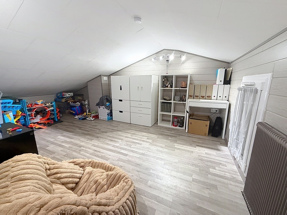 Pièce sous combles aménagée avec sol en parquet stratifié gris clair et plafond incliné recouvert de lambris gris. Rangements blancs IKEA, bibliothèque à cases, bureau blanc, radiateur à ailettes et jouets pour enfant installés sous la charpente apparente.