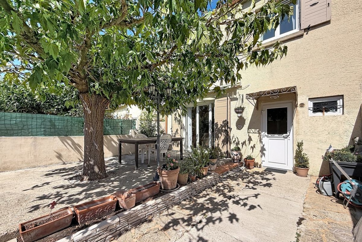 Jardin clos avec un grand arbre feuillu central projetant de l'ombre sur une terrasse en béton avec table et chaises blanches. Façade en enduit beige avec volets gris, bordure de jardinières en terre cuite alignées et plusieurs plantes en pots disposées le long de la maison.