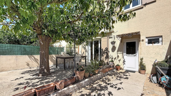 Jardin clos avec un grand arbre feuillu central projetant de l'ombre sur une terrasse en béton avec table et chaises blanches. Façade en enduit beige avec volets gris, bordure de jardinières en terre cuite alignées et plusieurs plantes en pots disposées le long de la maison.