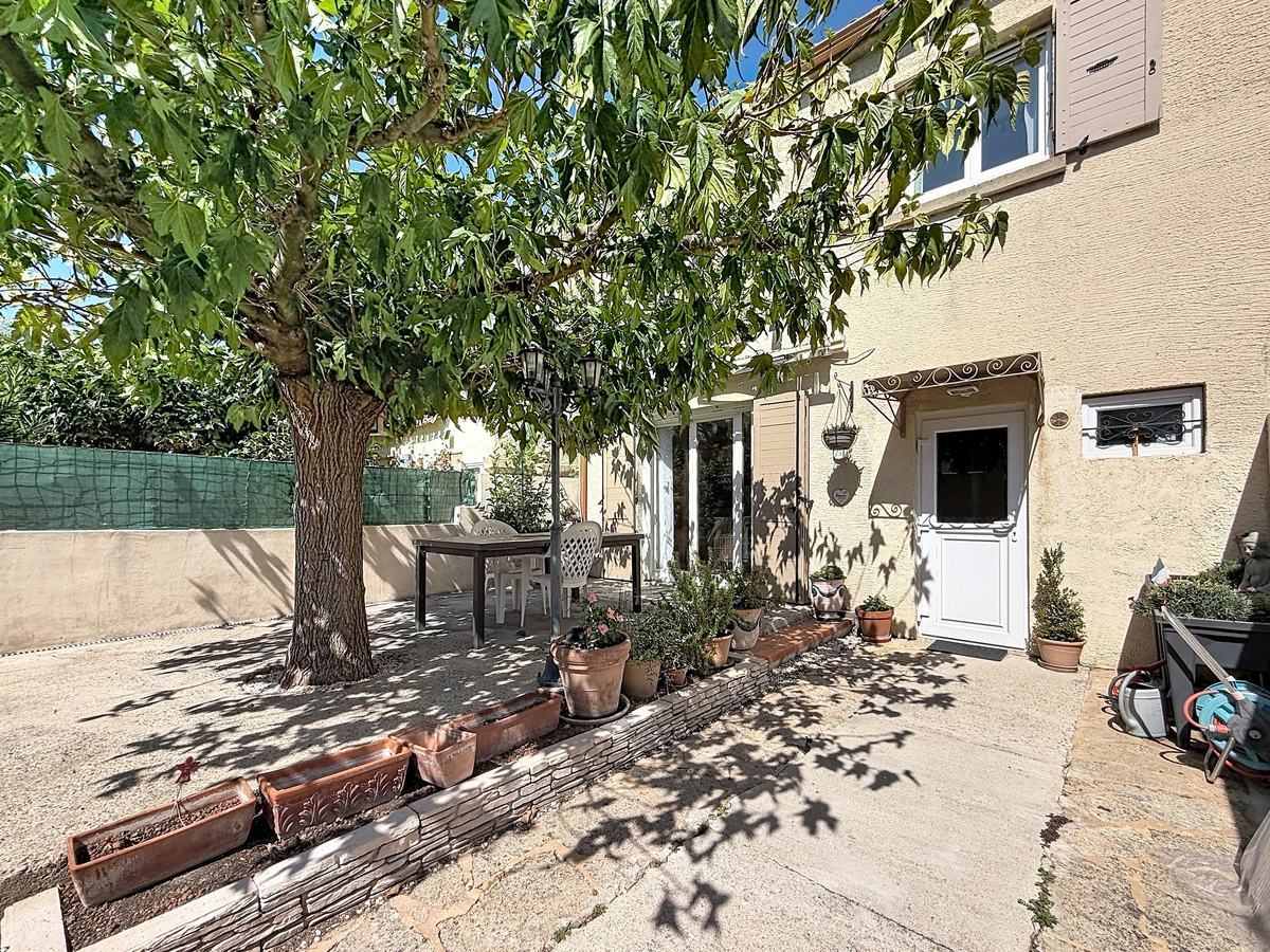 Jardin clos avec un grand arbre feuillu central projetant de l'ombre sur une terrasse en béton avec table et chaises blanches. Façade en enduit beige avec volets gris, bordure de jardinières en terre cuite alignées et plusieurs plantes en pots disposées le long de la maison.
