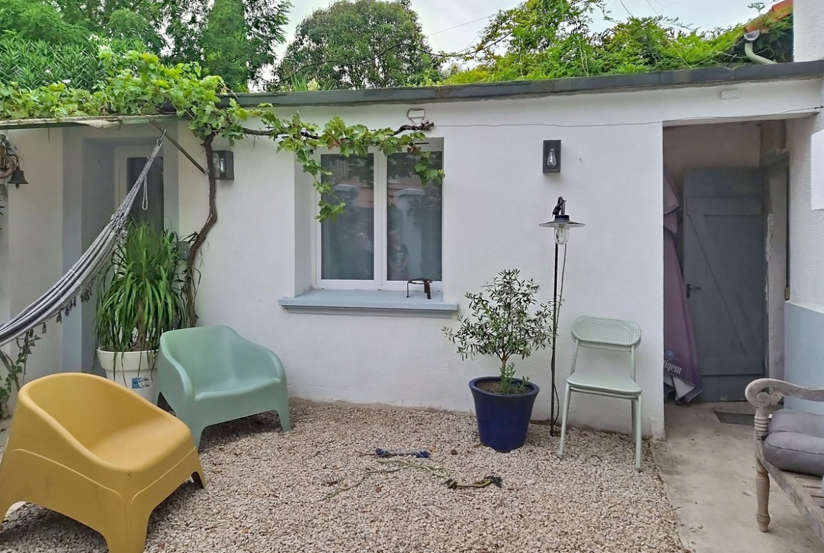 Cour extérieure recouverte de graviers clairs avec une dépendance blanche en fond, une vigne grimpante courant sur la façade et le toit végétalisé. Plusieurs chaises de jardin en plastique coloré (vert, jaune), un olivier en pot bleu et un hamac visible sur la gauche.