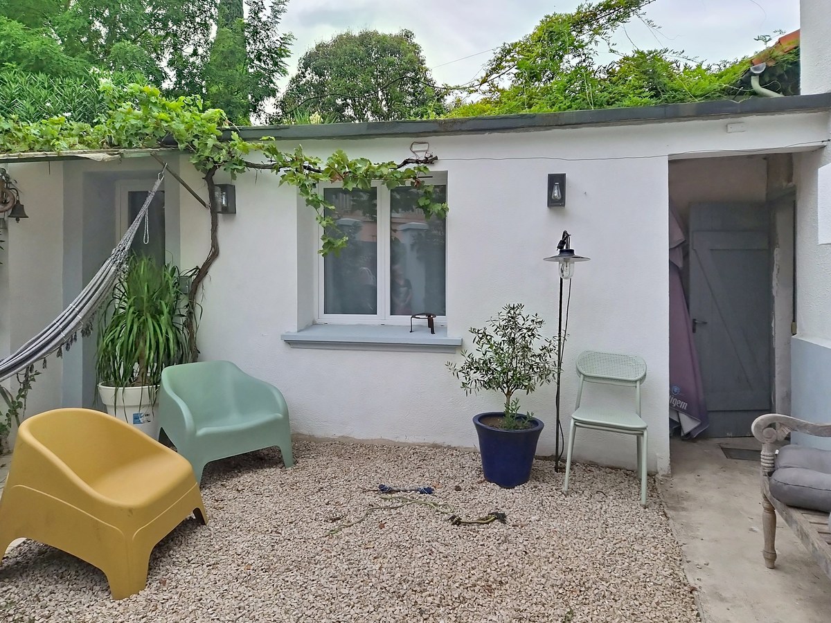 Cour extérieure recouverte de graviers clairs avec une dépendance blanche en fond, une vigne grimpante courant sur la façade et le toit végétalisé. Plusieurs chaises de jardin en plastique coloré (vert, jaune), un olivier en pot bleu et un hamac visible sur la gauche.