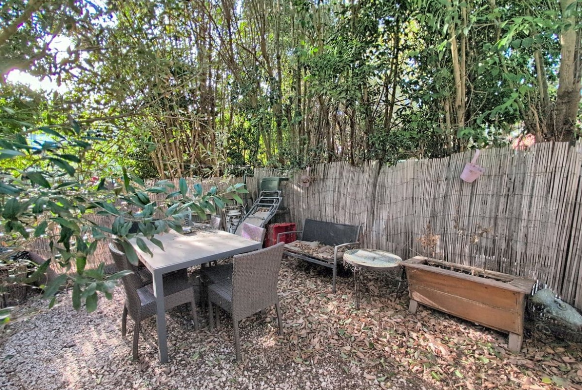 Espace extérieur privatif avec sol en graviers et table de jardin rectangulaire grise entourée de quatre chaises en résine tressée. Clôture en canisse naturelle délimitant le fond du jardin, avec végétation dense composée de bambous et arbustes à feuillage vert.