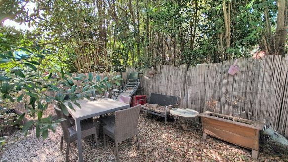 Espace extérieur privatif avec sol en graviers et table de jardin rectangulaire grise entourée de quatre chaises en résine tressée. Clôture en canisse naturelle délimitant le fond du jardin, avec végétation dense composée de bambous et arbustes à feuillage vert.