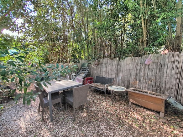 Espace extérieur privatif avec sol en graviers et table de jardin rectangulaire grise entourée de quatre chaises en résine tressée. Clôture en canisse naturelle délimitant le fond du jardin, avec végétation dense composée de bambous et arbustes à feuillage vert.