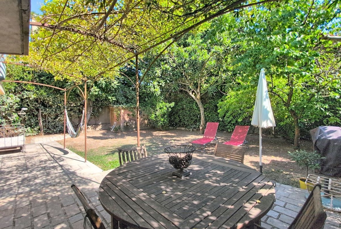 Terrasse dallée en pierre agrémentée d'une table de jardin ronde en teck et de chaises assorties, sous une pergola métallique avec glycine, entourée d'arbres méditerranéens matures dont un magnolia et des figuiers. Un hamac, deux chaises longues rouges et un parasol blanc sont disposés dans le jardin en fond de parcelle.