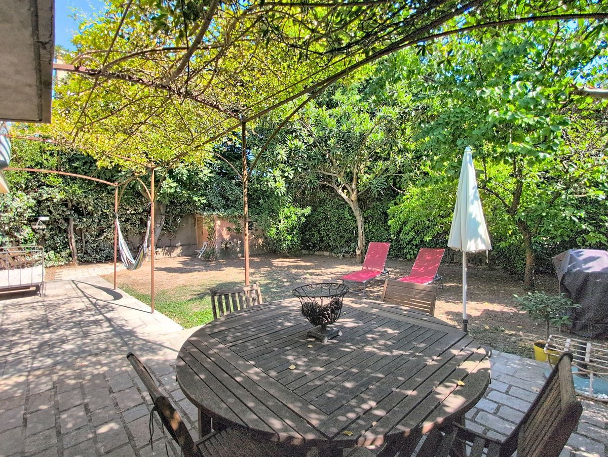 Terrasse dallée en pierre agrémentée d'une table de jardin ronde en teck et de chaises assorties, sous une pergola métallique avec glycine, entourée d'arbres méditerranéens matures dont un magnolia et des figuiers. Un hamac, deux chaises longues rouges et un parasol blanc sont disposés dans le jardin en fond de parcelle.