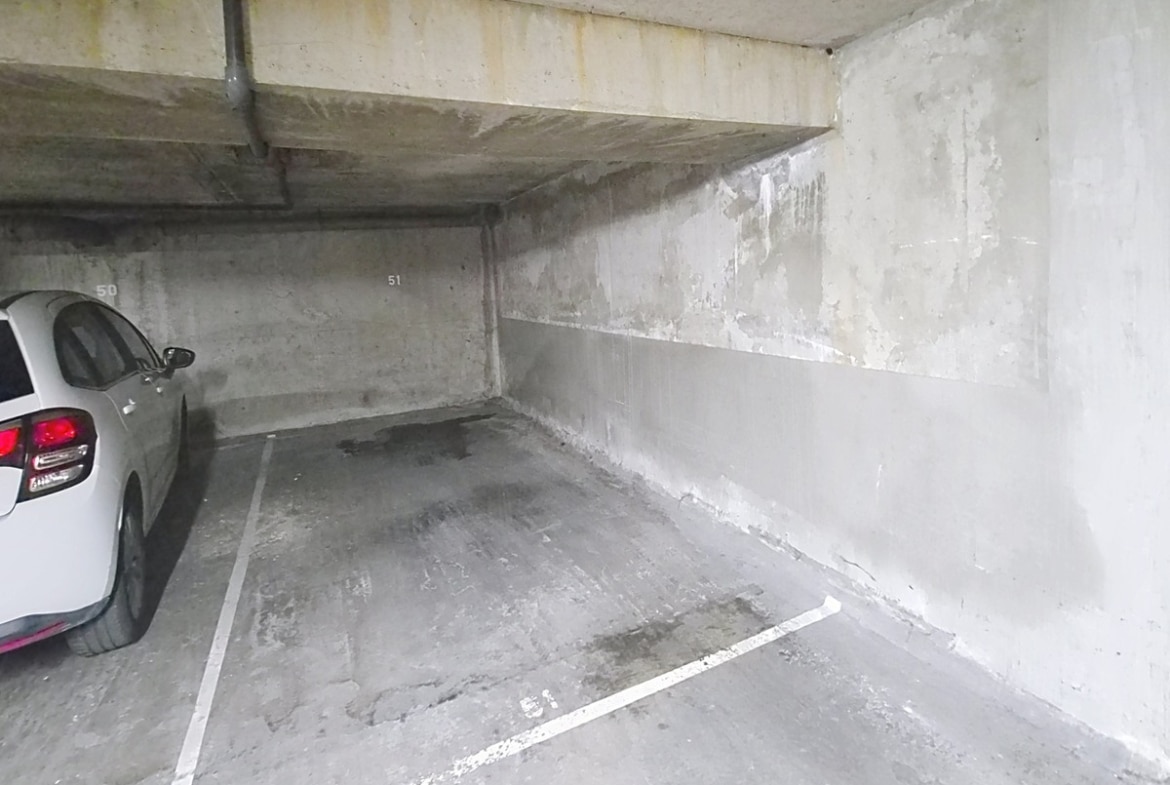 Parking souterrain en béton brut avec une place numérotée 51 délimitée par des marquages blancs au sol, entourée de murs en béton gris et d'un plafond bas avec des tuyaux apparents. Une voiture blanche est garée sur la place adjacente numérotée 50, à gauche du cadre.