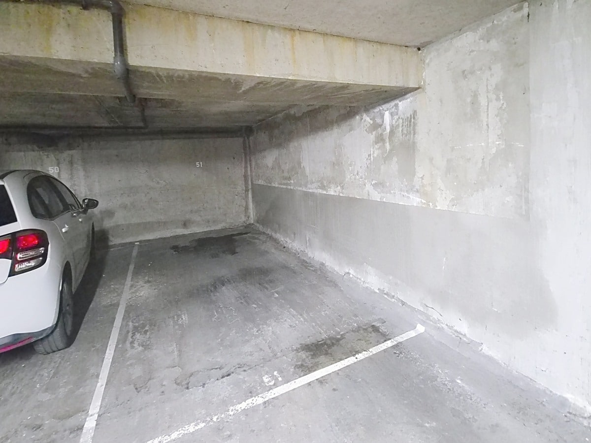 Parking souterrain en béton brut avec une place numérotée 51 délimitée par des marquages blancs au sol, entourée de murs en béton gris et d'un plafond bas avec des tuyaux apparents. Une voiture blanche est garée sur la place adjacente numérotée 50, à gauche du cadre.