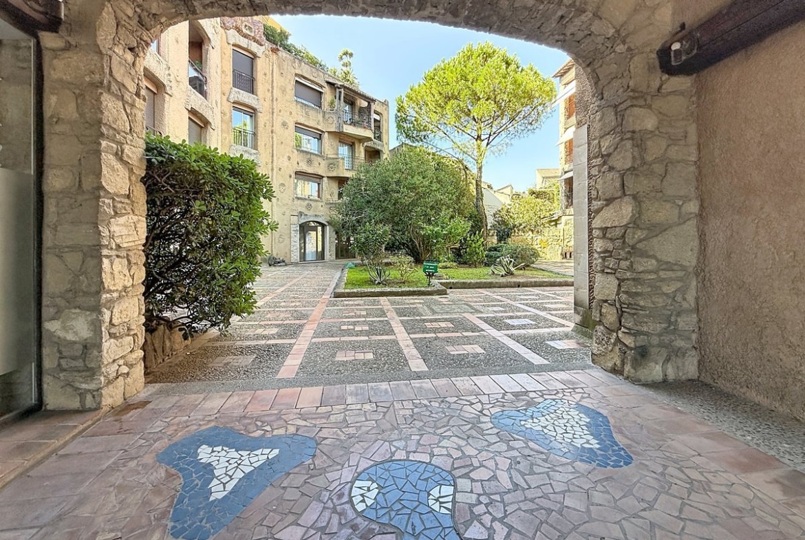 Passage voûté en pierre de taille donnant accès à une cour intérieure pavée avec un sol décoré d'une mosaïque en carrelage bleu et gris en forme de personnage, entouré d'espaces verts soignés avec arbustes et un grand pin parasol. La résidence en pierre apparente de plusieurs étages est visible en arrière-plan avec des balcons et des volets roulants.
