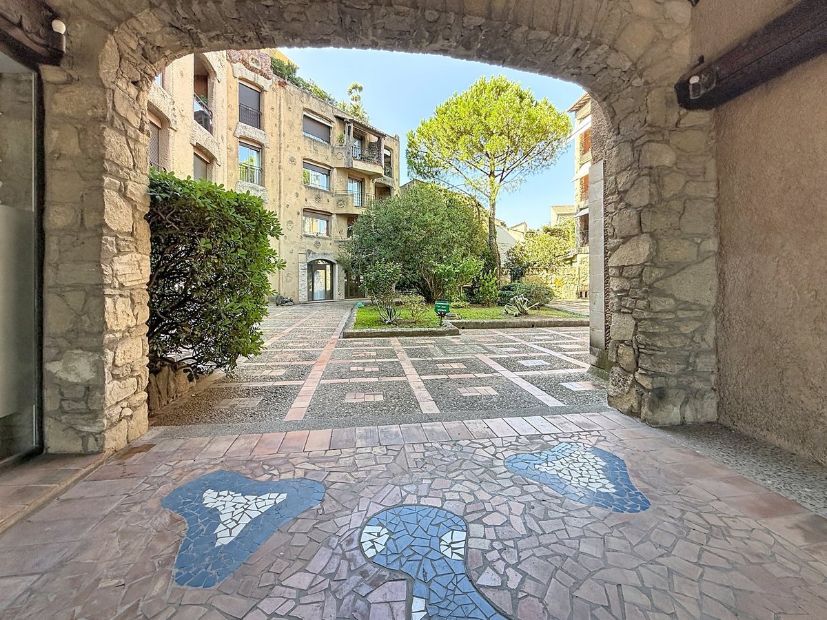 Passage voûté en pierre de taille donnant accès à une cour intérieure pavée avec un sol décoré d'une mosaïque en carrelage bleu et gris en forme de personnage, entouré d'espaces verts soignés avec arbustes et un grand pin parasol. La résidence en pierre apparente de plusieurs étages est visible en arrière-plan avec des balcons et des volets roulants.