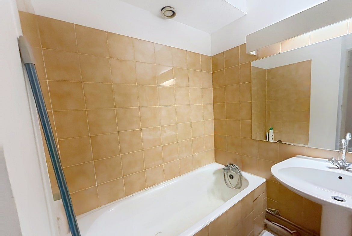 Salle de bain entièrement carrelée de grands carreaux beige-ocre brillants, équipée d'une baignoire encastrée blanche avec mitigeur chromé et d'un lavabo vasque blanc sur meuble bois arrondi. Un miroir rectangulaire sans cadre est fixé au-dessus du lavabo, et une ventilation mécanique circulaire est visible au plafond blanc.